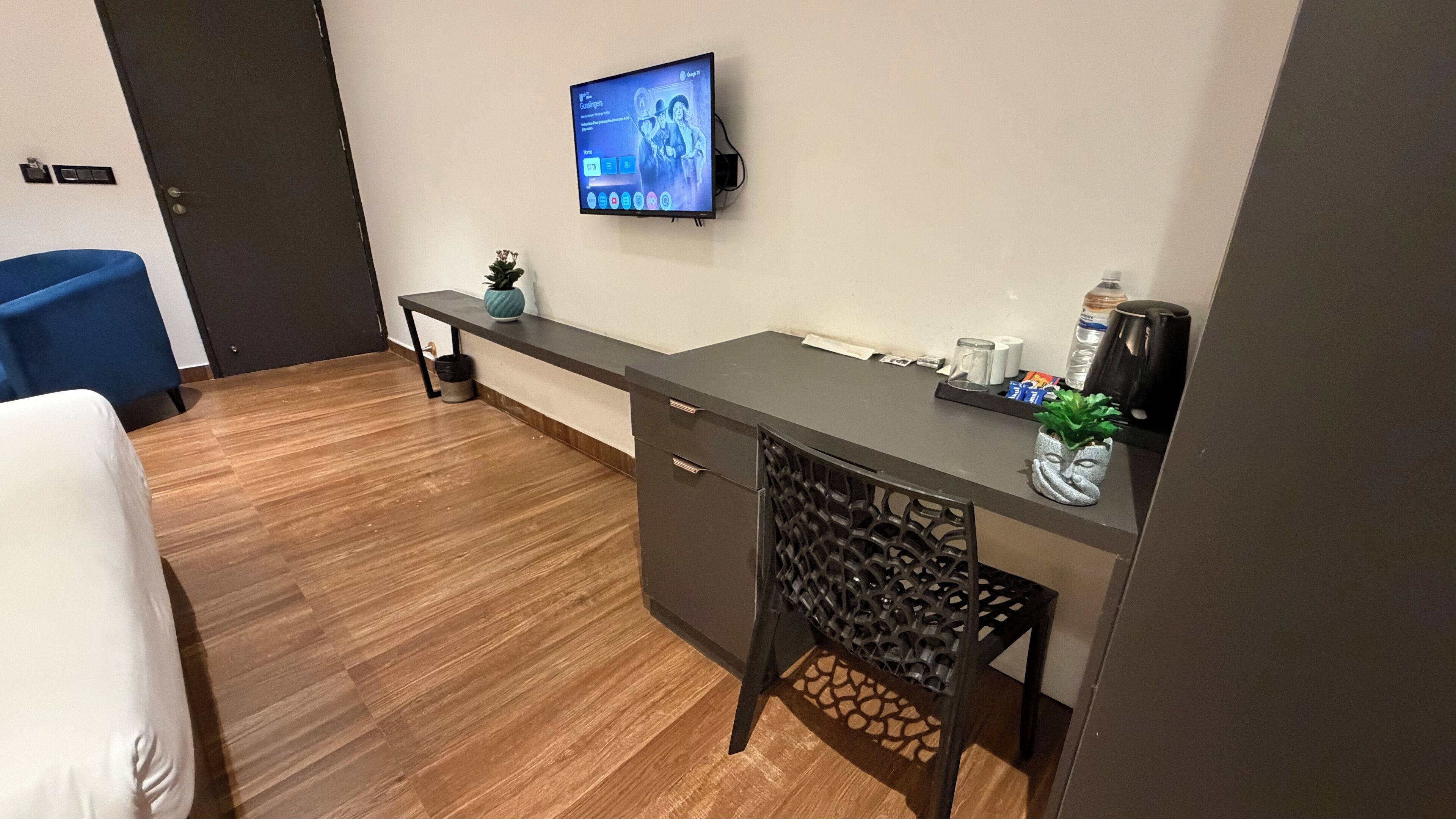 Una habitación de hotel moderna con piso de madera, un escritorio con televisión de pantalla plana, una silla azul y una cama con ropa de cama blanca.