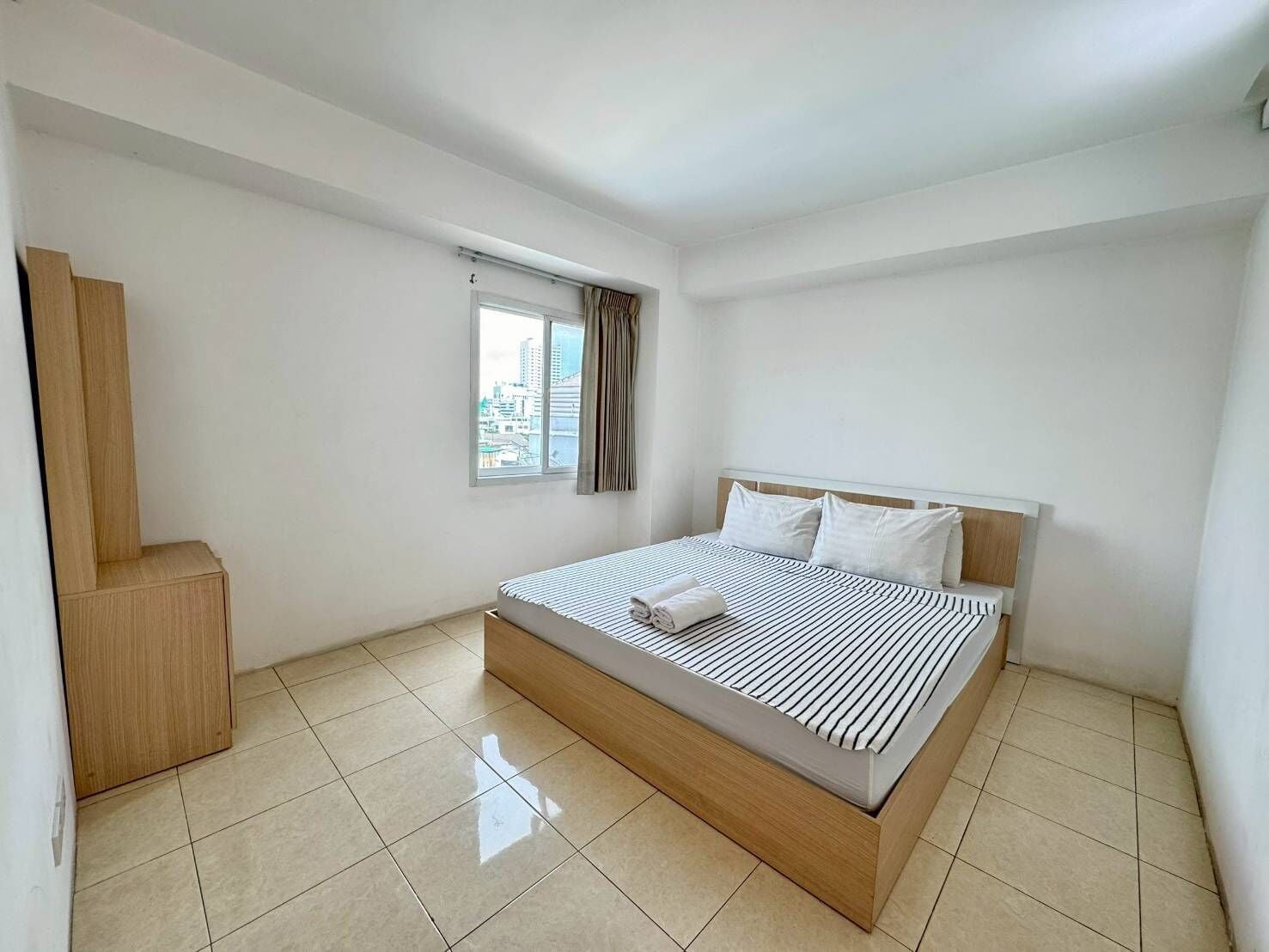 A modern hotel room with a large bed, a wooden wardrobe, a window with curtains, and a city view.