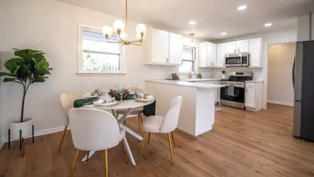 A modern kitchen with white cabinets, a central island, and a dining area with white chairs and a chandelier.