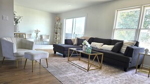 A modern living room with a dark grey sofa, white armchairs, and a glass coffee table. There is a bookshelf and a vase with flowers on a side table.