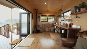 A cozy cabin interior with a wooden deck, a kitchen area, and a large window overlooking a scenic view. - Sunsets, outdoor bath, mountain views, fire pit (Marotiri)