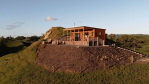 A modern wooden house with a covered porch and a stone staircase leading up to it, set in a hilly landscape. - Sunsets, outdoor bath, mountain views, fire pit (Marotiri)