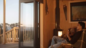 A person sitting on a deck chair by a glass door, holding a guitar, with a wood-burning stove in the background. - Sunsets, outdoor bath, mountain views, fire pit (Marotiri)