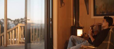A person sitting on a deck chair by a glass door, holding a guitar, with a wood-burning stove in the background.