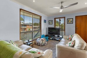 A living room with a TV, two sofas, a ceiling fan, and large windows with blinds. - Welcome to "Shore Beats Work!" (Gympie)