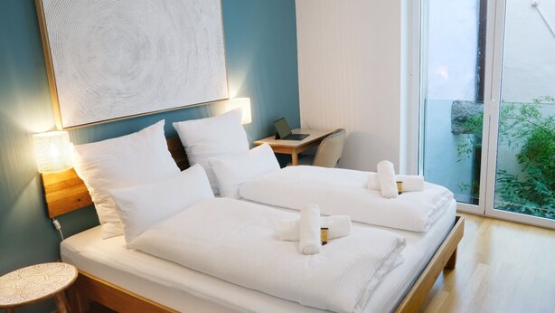 A hotel room with two beds, a small table, a chair, and a large window. - Apartment Lauben- Alpstay (Merano)