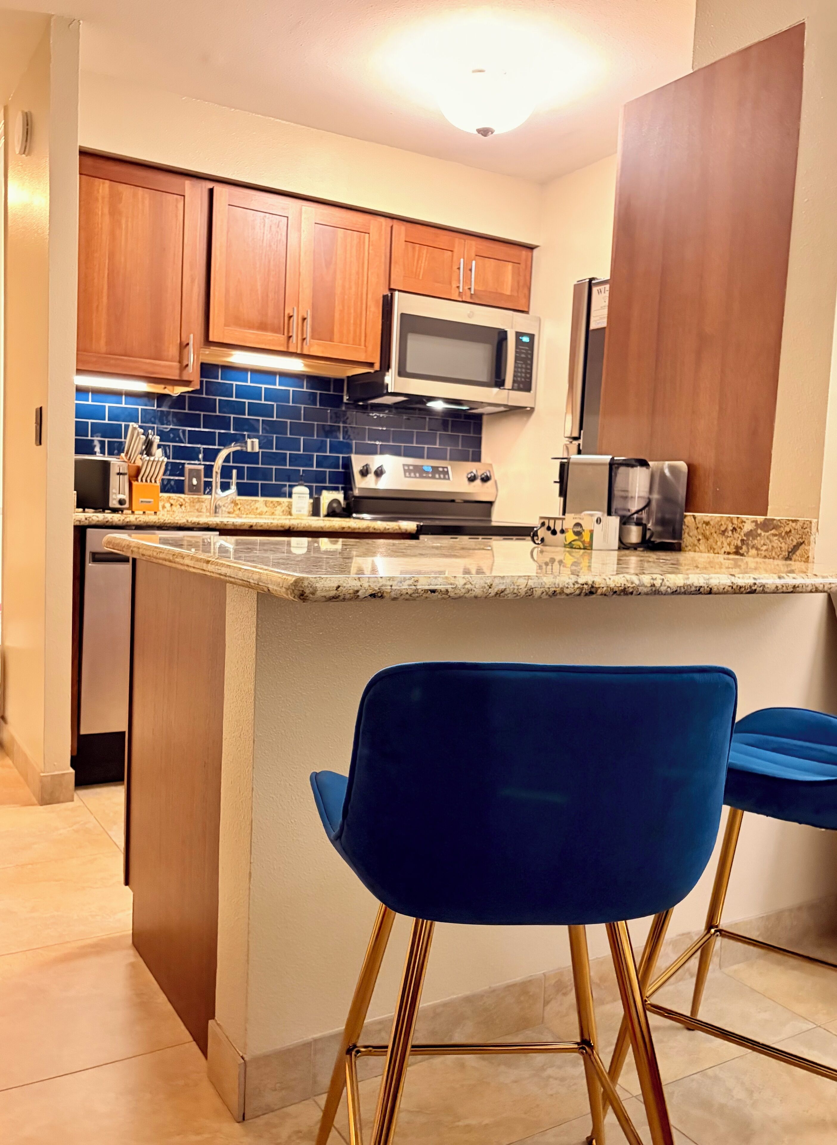 A kitchen with wooden cabinets, a microwave, a stove, and a sink. There are two blue chairs with gold legs.
