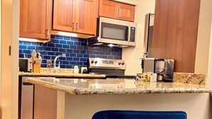 A kitchen with wooden cabinets, a microwave, a stove, and a sink. There are two blue chairs with gold legs. - Guest Favorite: Beachfront Studio Turtles, Monk Seals, Surf & Mountain View (Lahaina)