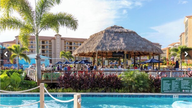 A resort pool area with a thatched-roof pavilion, lounge chairs, and a pool with a depth marker. - Spacious 3-bedroom condo with AC in beautiful Orlando (Orlando)