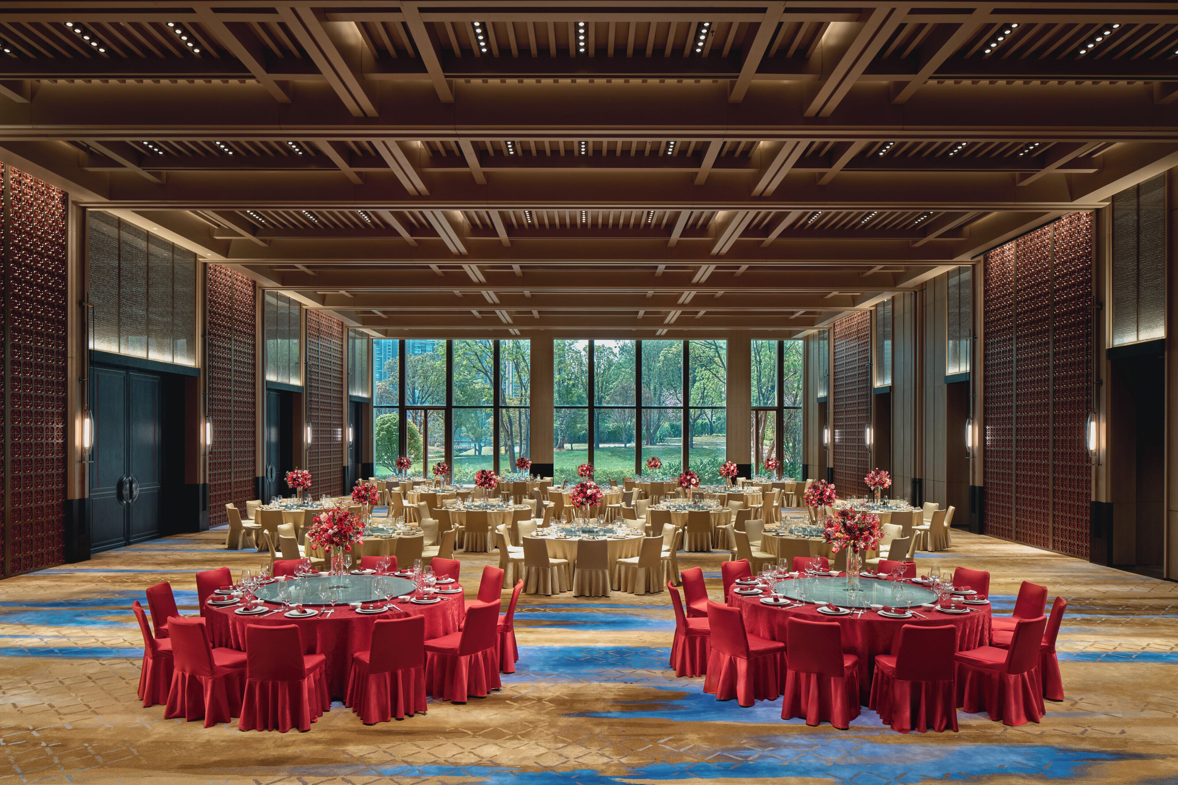 A large banquet hall with multiple round tables, red chairs, and a view of greenery through large windows.