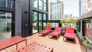 A rooftop terrace with red lounge chairs, a wooden table, and potted plants. - Iconic Queensbridge New Designer Suite with Heated Pool, Gym, Sauna, and Steam (Melbourne)
