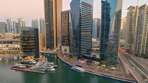 Un paysage urbain au bord de l’eau, avec des immeubles modernes de grande hauteur, une marina remplie de bateaux et une promenade piétonne longeant la rivière.