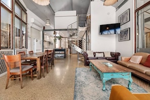A spacious living area with a dining table, sofas, a staircase, and a TV mounted on the wall.