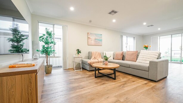 Uma sala de estar ampla com sofá, mesa de centro e pequena mesa lateral. Há uma planta em vaso e um quadro na parede.