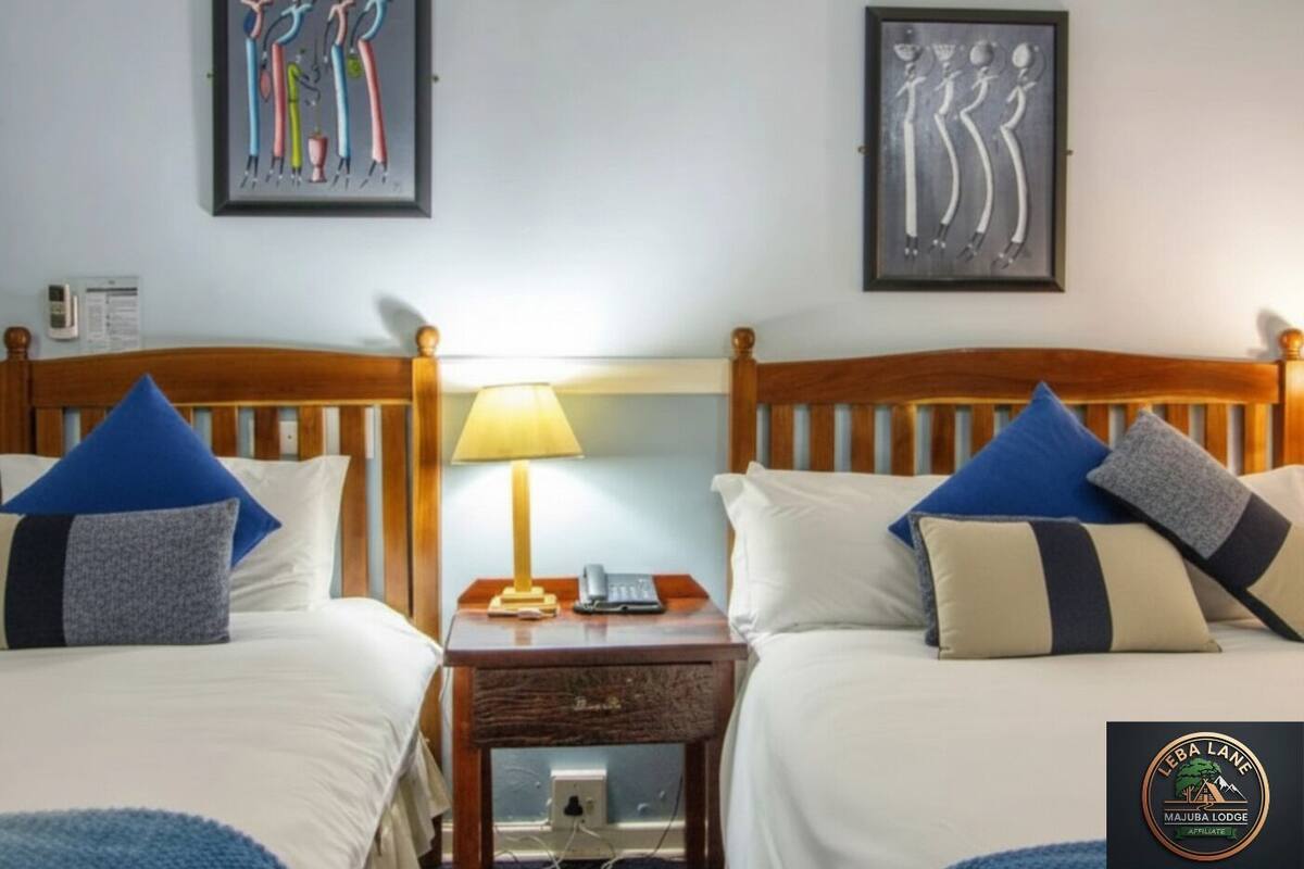 A hotel room with two beds, each with blue and grey pillows, a wooden headboard, a bedside table with a lamp, and artwork on the walls.