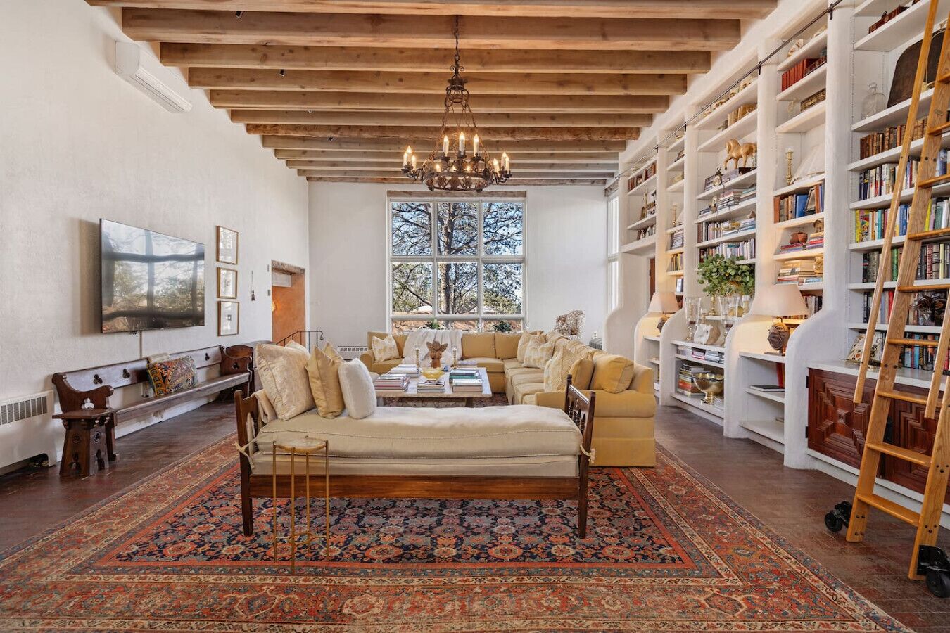 A spacious living room with a large sofa set, a wooden coffee table, and a patterned rug. There is a bookshelf filled with books and decorative items, and a chandelier hangs from the ceiling.