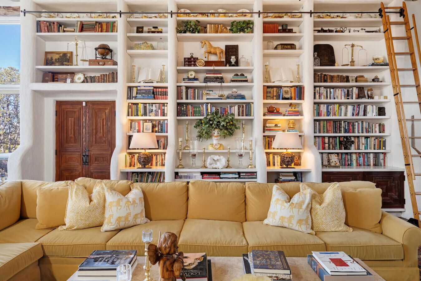 Una habitación con un sofá beige grande, múltiples estanterías llenas de libros y objetos decorativos, y una escalera de madera apoyada contra la pared.