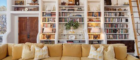 Een kamer met een grote beige bank, meerdere boekenkasten vol boeken en decoratieve objecten, en een houten ladder die tegen de muur staat.