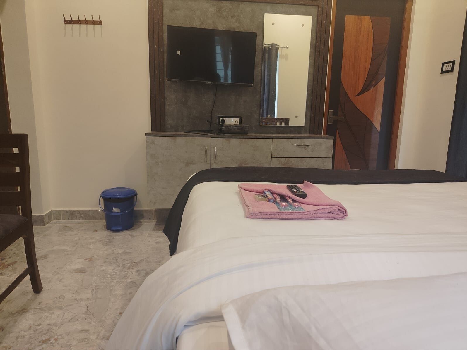 Ein Hotelzimmer mit einem Bett, einem an der Wand befestigten Fernseher, einem Holzschrank und einem blauen Eimer auf dem Boden.