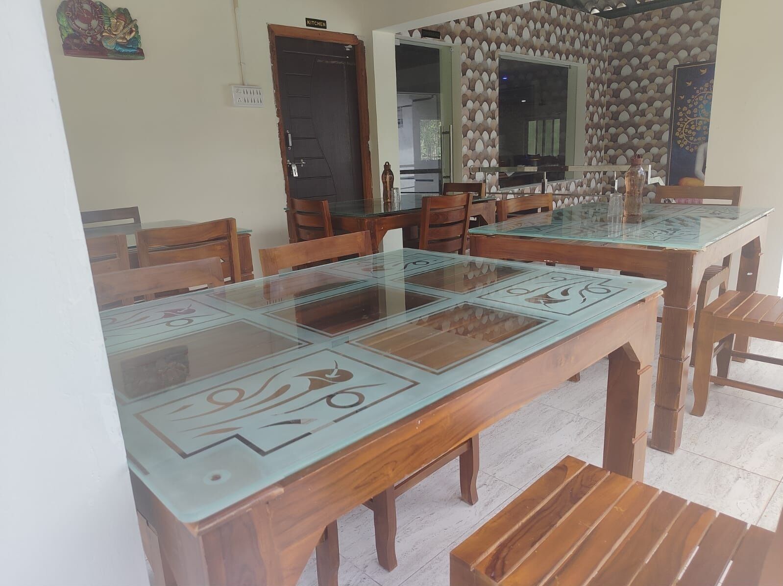 A dining area with a long wooden table and chairs, a kitchen visible through an open door, and a patterned wall.
