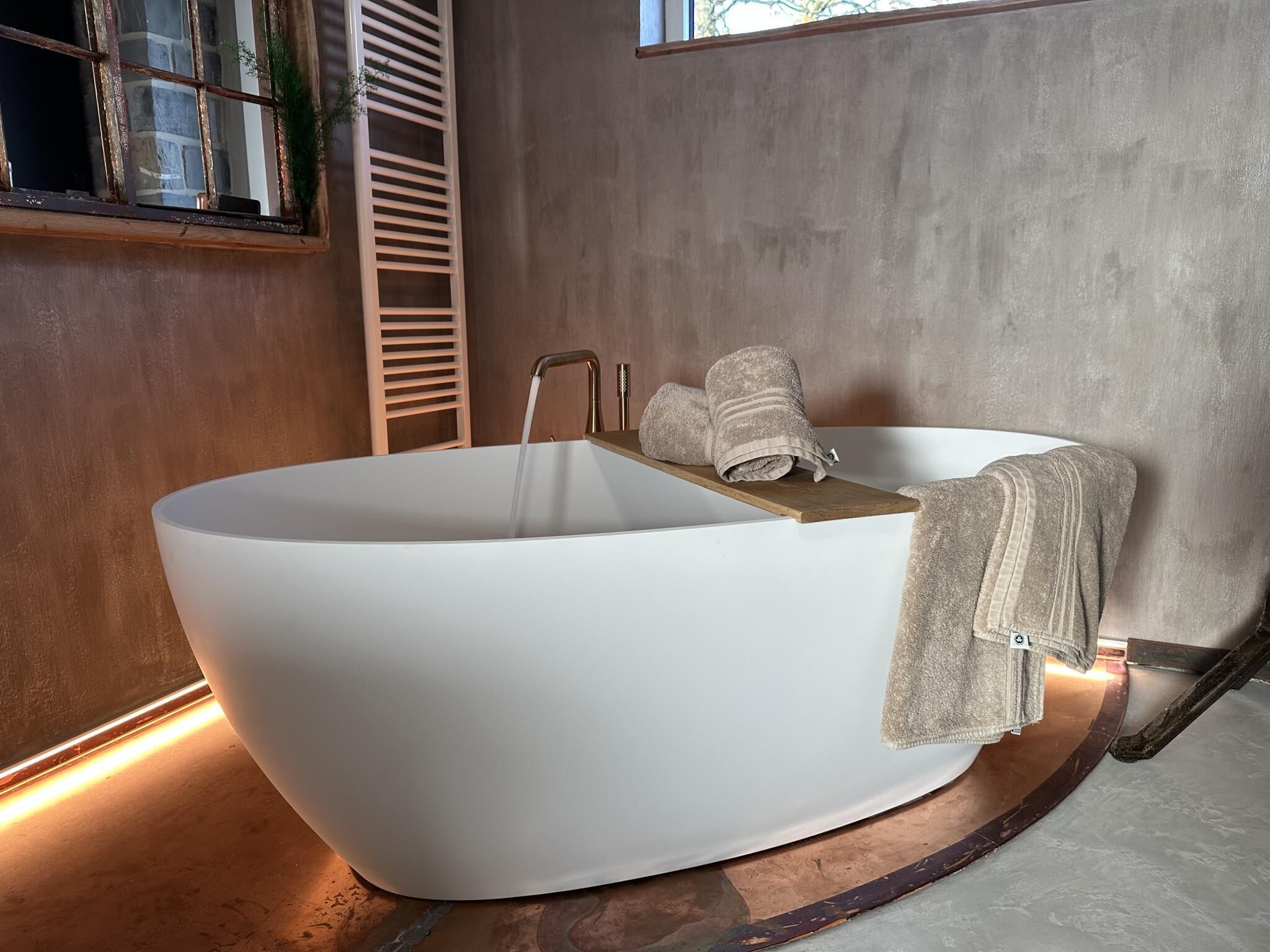 Un baño moderno con una bañera independiente, un estante de madera con toallas y una ventana con vistas a árboles.