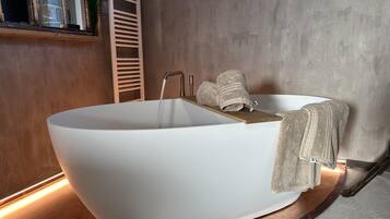 Un baño moderno con una bañera independiente, un estante de madera con toallas y una ventana con vistas a árboles.