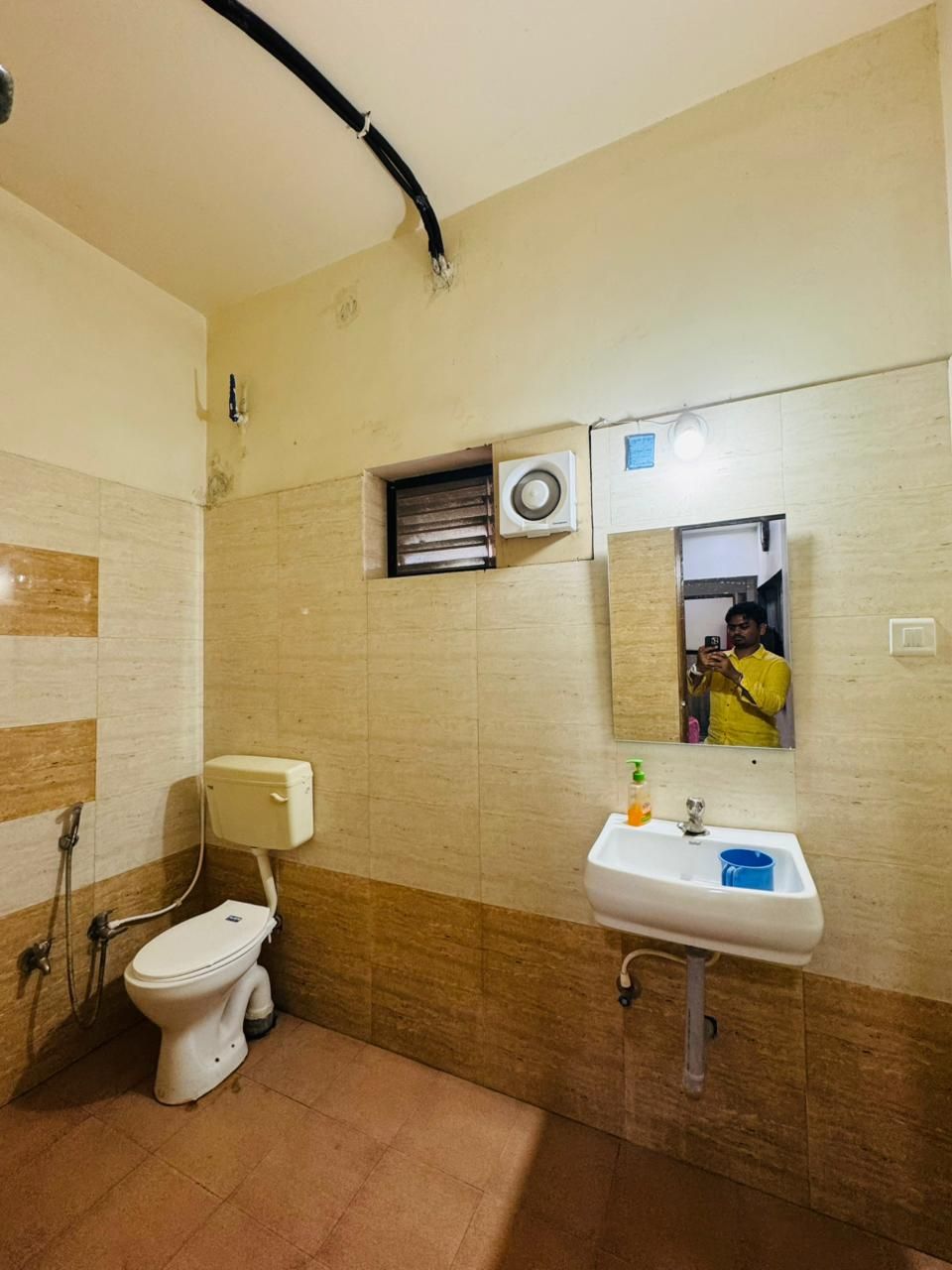 A bathroom with a toilet, sink, mirror, and a small window with an air conditioning unit.