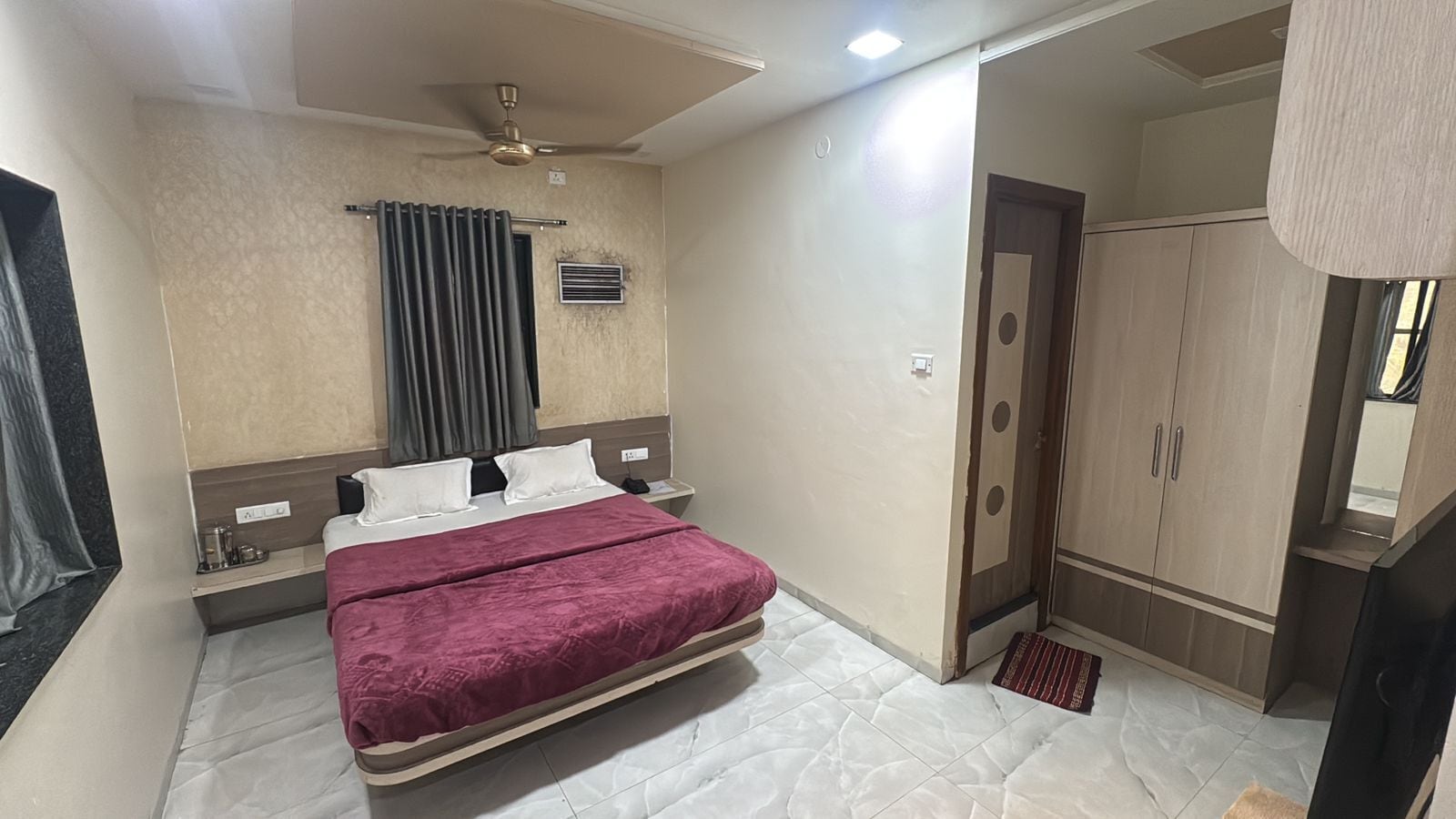 A hotel room with a bed, a ceiling fan, a wardrobe, and a small table with a lamp.