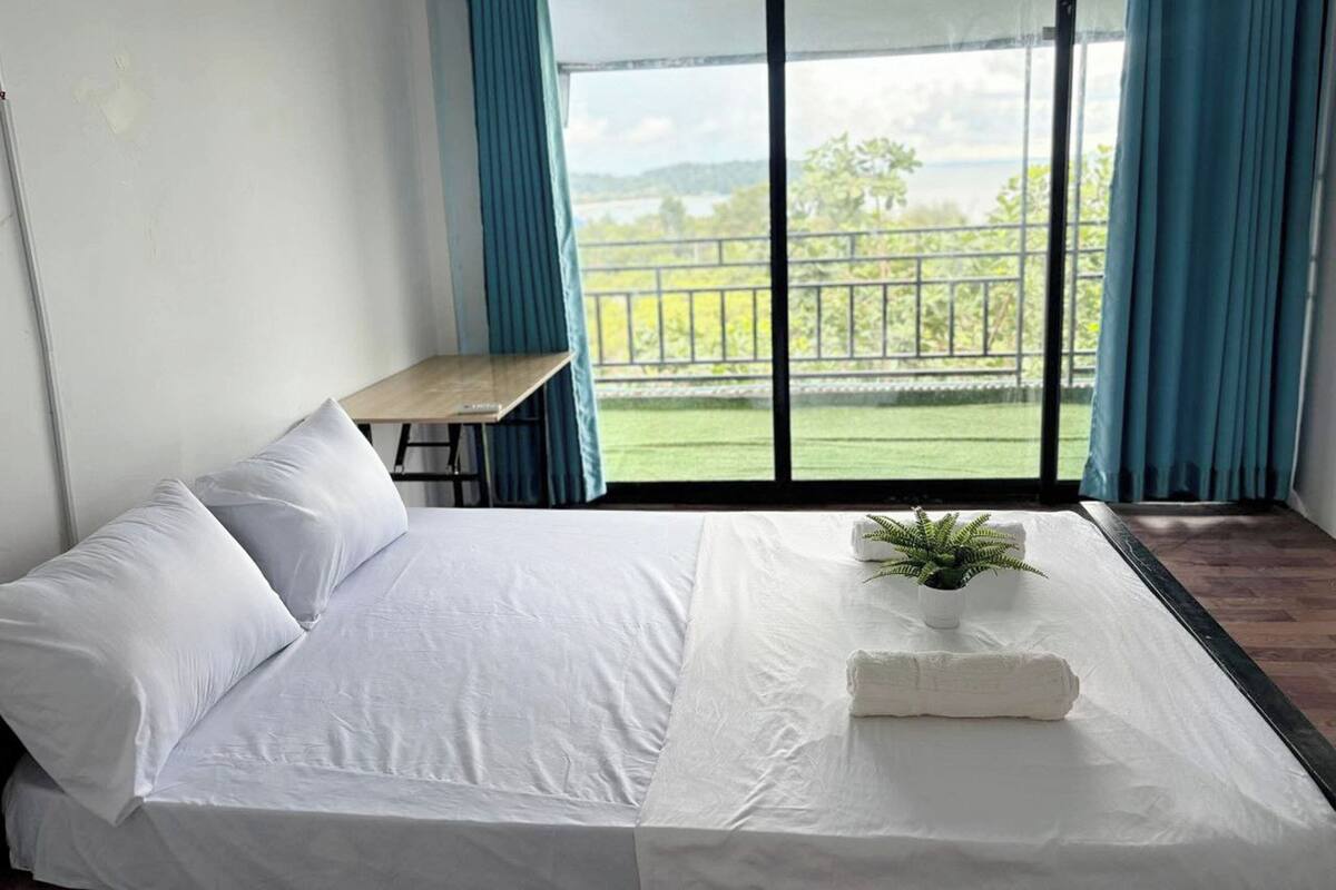 A bedroom with a bed, a small table, and a view of a green landscape through large windows.