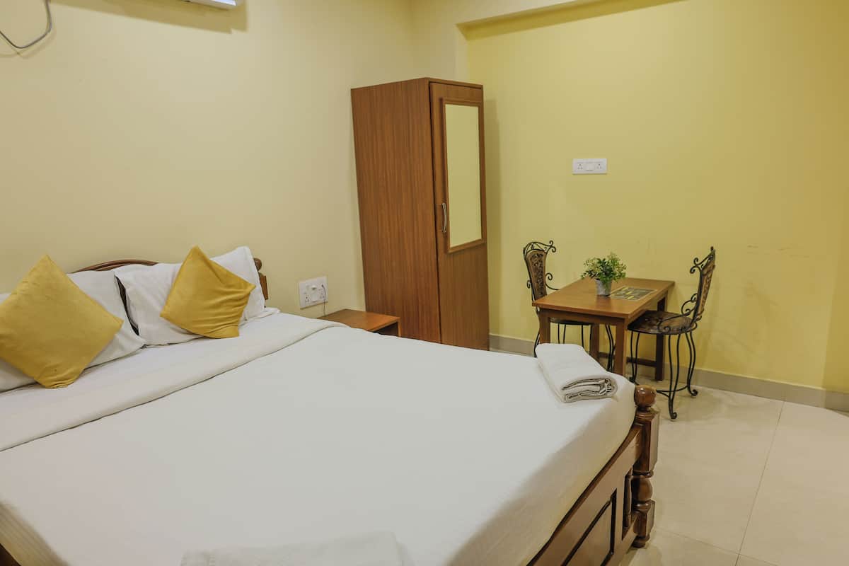 A neatly made bed with white and yellow pillows, a wooden wardrobe, a small wooden table with a chair, and a wall socket.