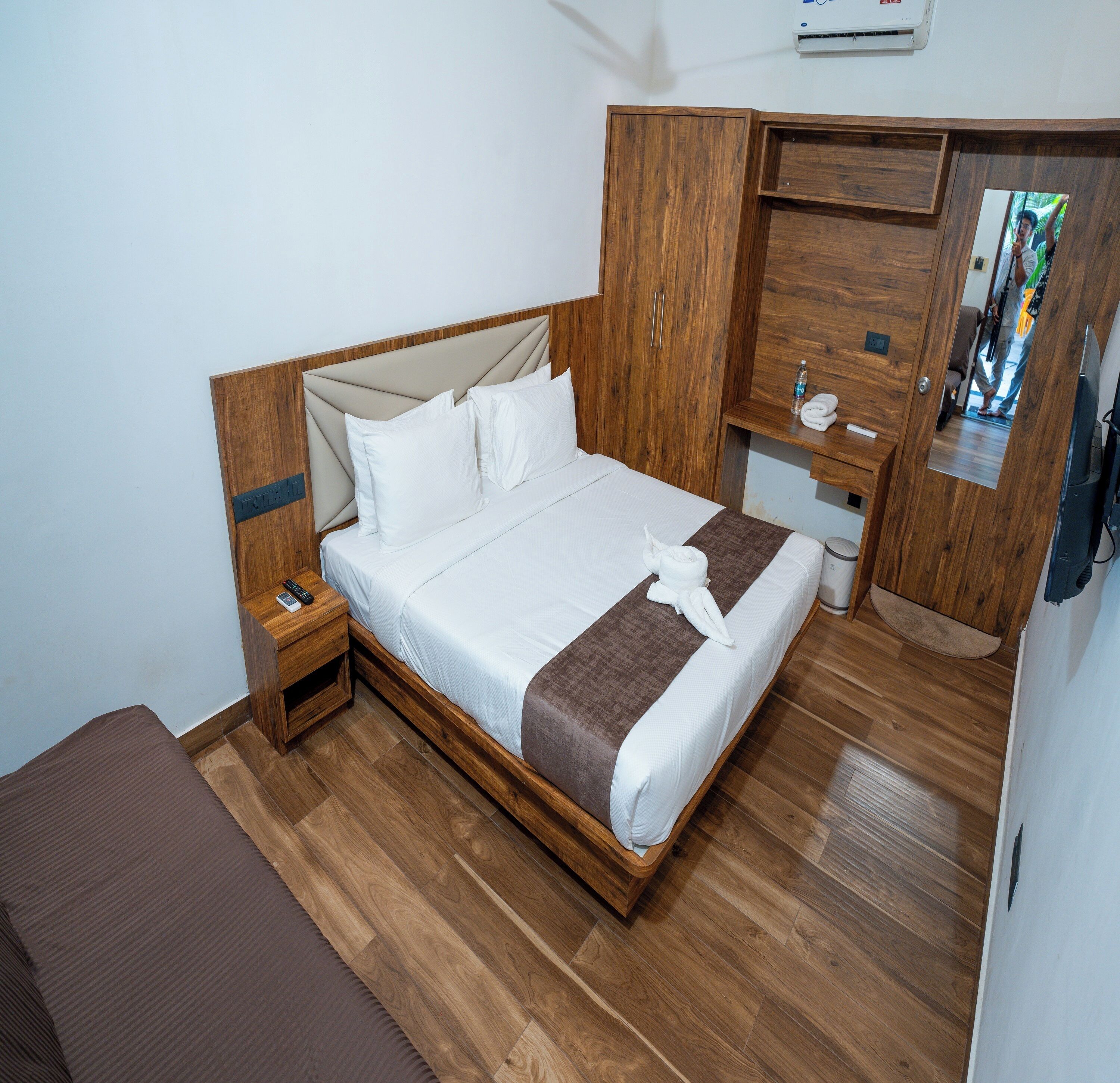 Habitación de hotel ordenada con una cama de madera, una mesita de noche con control remoto, un pequeño armario de madera y un espejo.