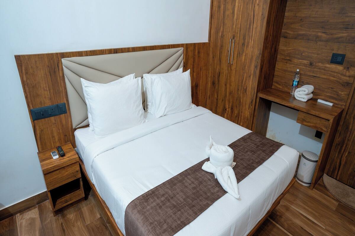 Een net opgemaakt bed met witte lakens, een bruine decoratieve plaid en een handdoek gevouwen op het bed. Er staat een houten nachtkastje met een afstandsbediening en een klein lade. Een houten garderobe met spiegel is op de achtergrond zichtbaar.