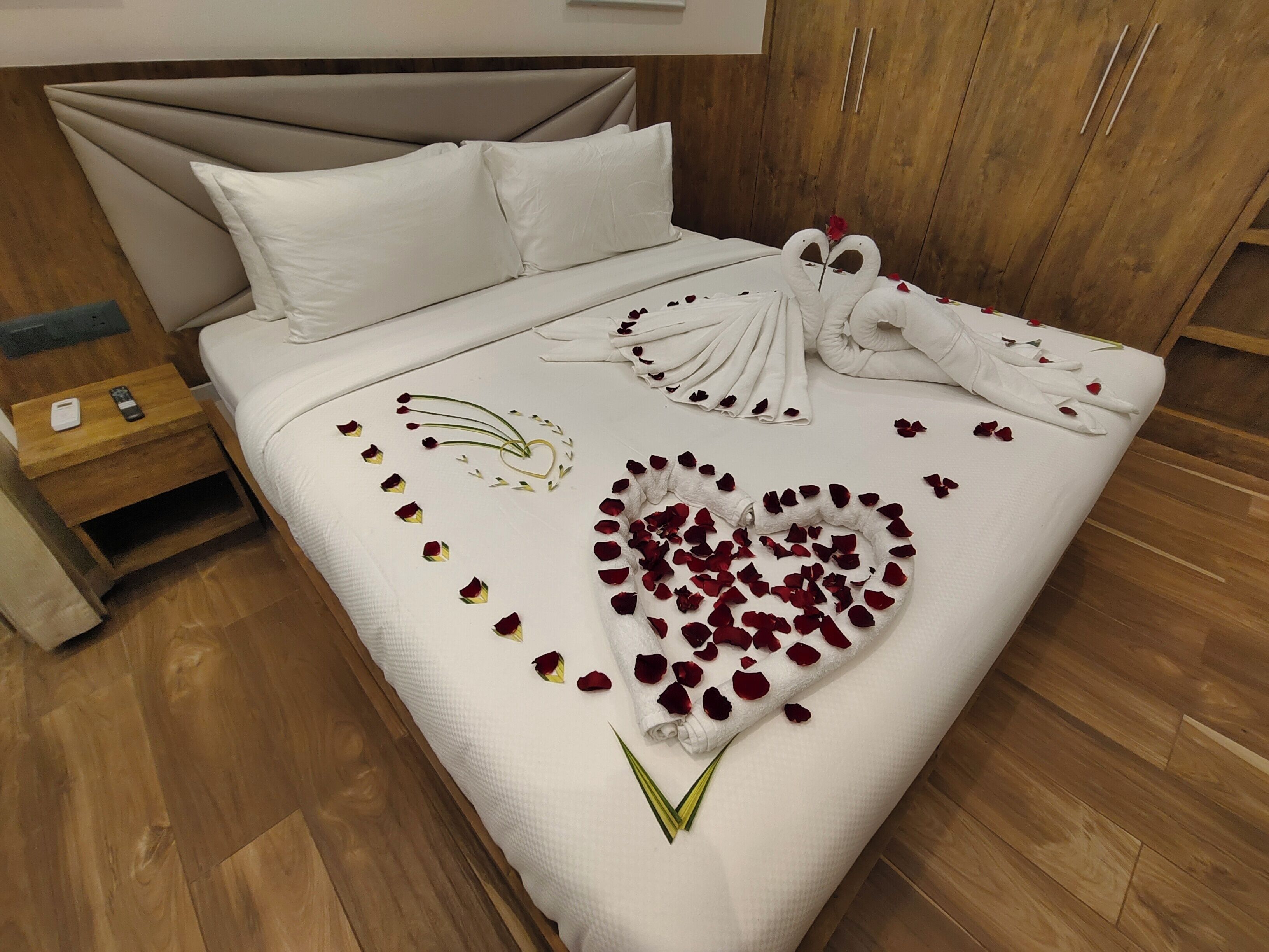 A neatly made bed with a heart-shaped arrangement of rose petals and a towel sculpture.