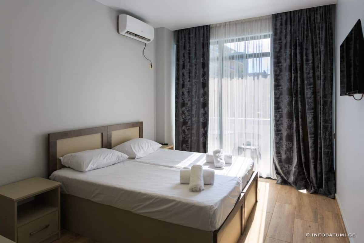 A hotel room with two beds, a bedside table, an air conditioning unit, a TV, and a large window with curtains.