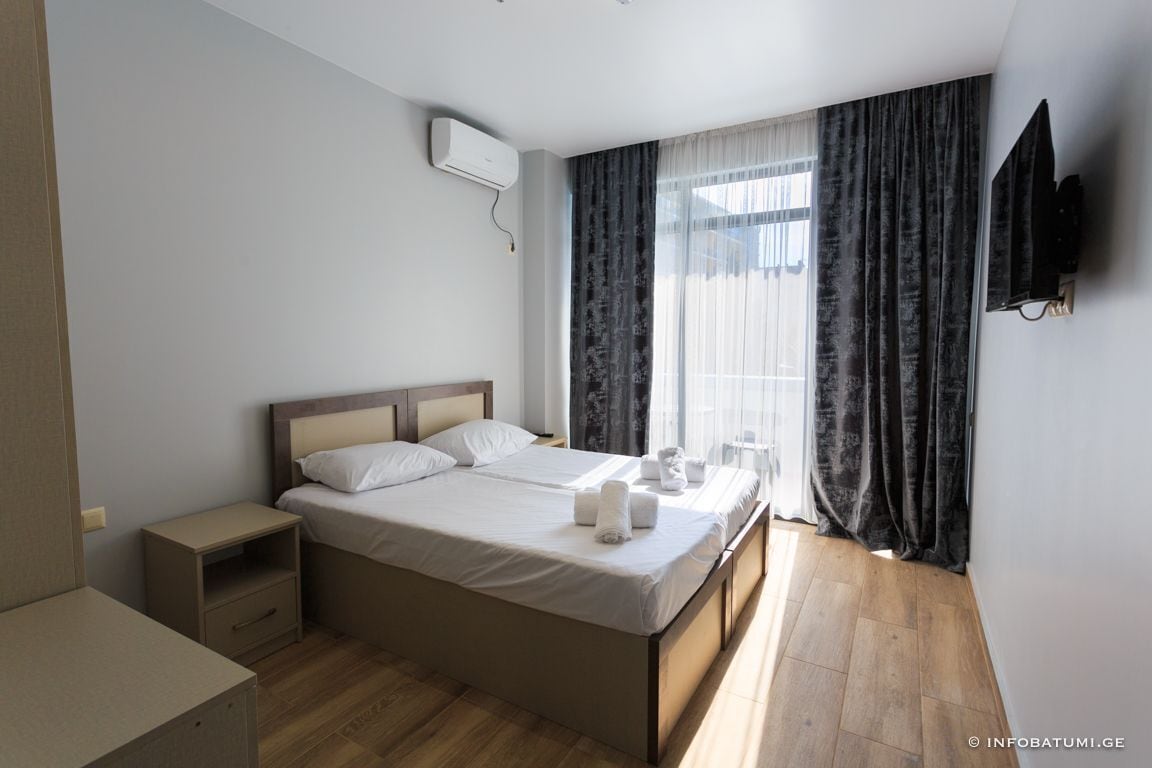 A double bed with a wooden headboard, a bedside table, and a small drawer unit. There is a window with sheer curtains and a dark drape, and a flat-screen TV mounted on the wall.