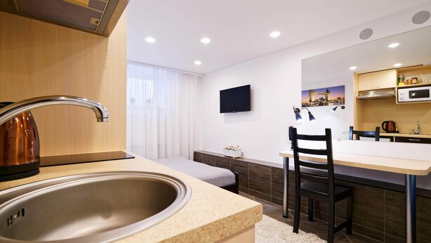 A modern kitchen with a sink, stove, and microwave. There is a dining area with a chair and a table. A TV is mounted on the wall above the dining area. - Compact Romantic Studio for couple (located in semi semi-basement) (Riga)
