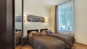 Hotelkamer met twee bedden, een grote spiegel, een stadslandschap schilderij, een raam met gordijnen en een nachtkastje met een lamp.