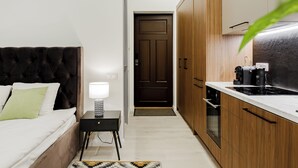 Ein kleines Hotelzimmer mit Bett, Nachttisch, Lampe und einer Küchenzeile inklusive Herd und Kaffeemaschine.