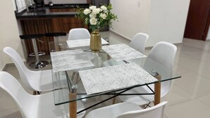 A modern dining area with a glass table and white chairs, a vase with white flowers, and a kitchen in the background. - Spacious accommodation with excellent location (Mazatlán)