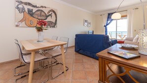 A dining area with a table and chairs, a sofa, a lamp, and a painting on the wall. - Casa da Gina Apartamento T2 com BBQ (Tavira)