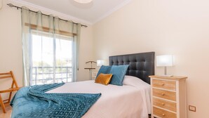 A bedroom with a bed, bedside tables, a lamp, a chair, and a window with curtains. - Casa da Gina Apartamento T2 com BBQ (Tavira)