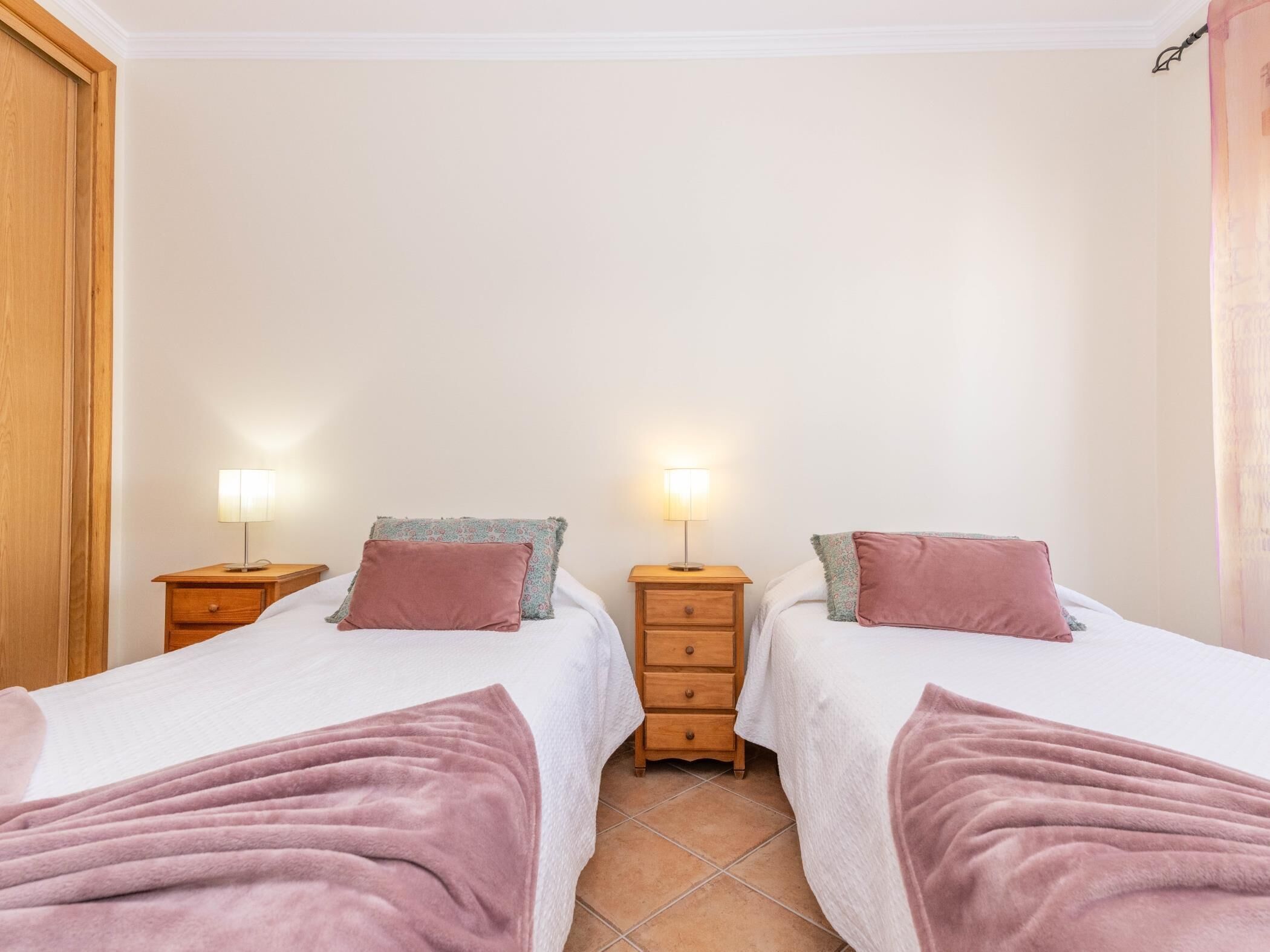 A double room with two beds, each with a bedside table and lamp, a wooden wardrobe, and a tiled floor.