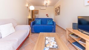 A living room with a sofa, a coffee table with seashells, a TV, and a bookshelf. - Casa da Gina Apartamento T2 com BBQ (Tavira)