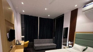 A hotel room with a bed, a desk, a chair, a sofa, and a ceiling fan. - Hotel Grand Soma (Nagpur)