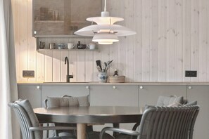 Un coin repas comprenant une table ronde, des chaises, un luminaire suspendu et un comptoir de cuisine avec des ustensiles et une cafetière.