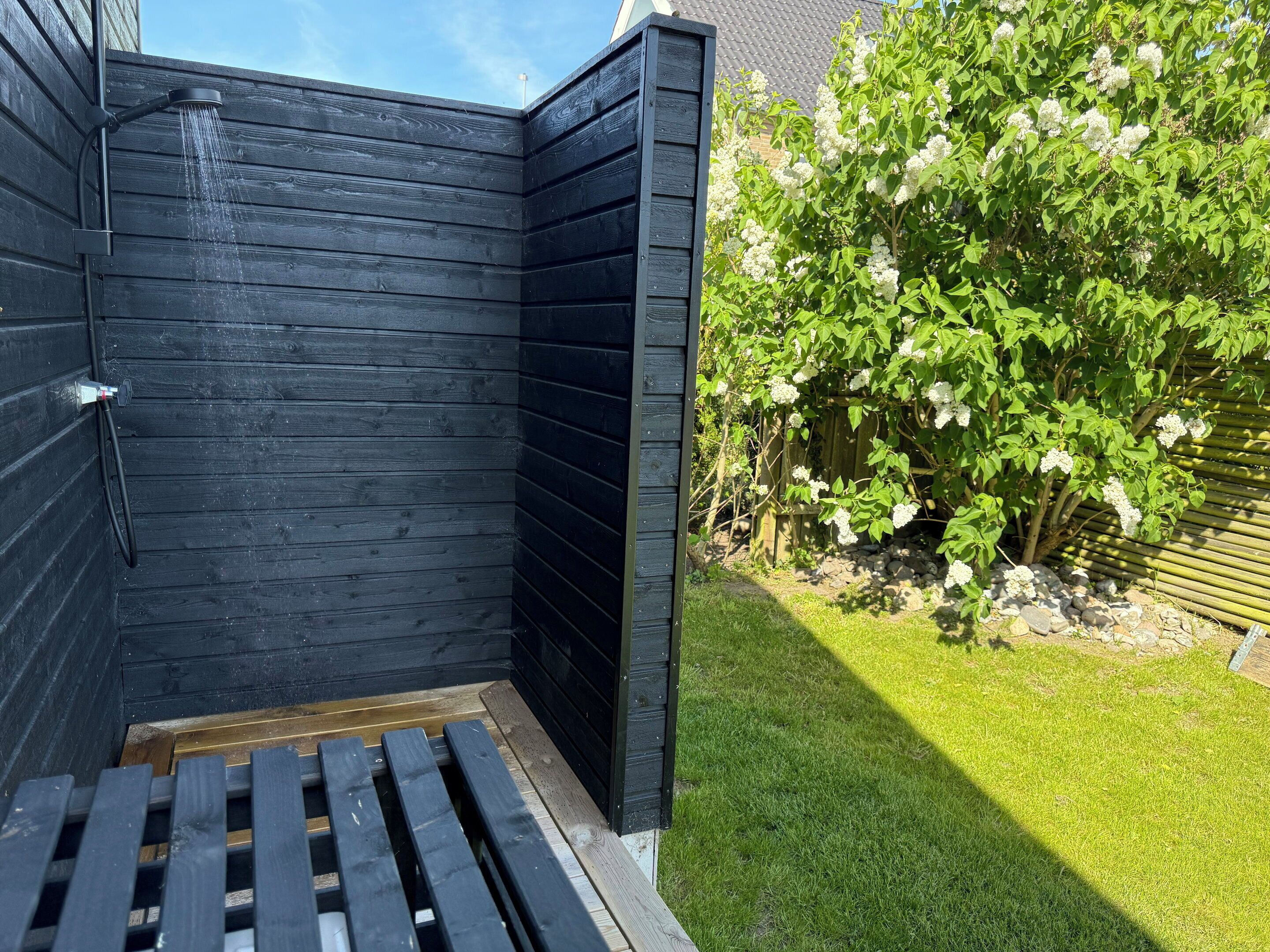 A wooden outdoor shower area with a wooden deck and a garden with white flowering bushes.