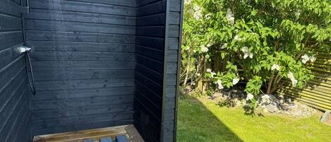 A wooden outdoor shower area with a wooden deck and a garden with white flowering bushes.
