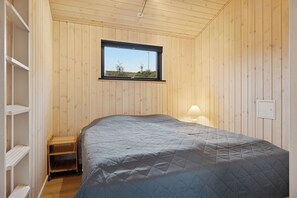A bedroom with a bed, a window, wooden walls, a shelf, and a lamp. - Charming newly built cottage 300 m from beach (Karrebæksminde)
