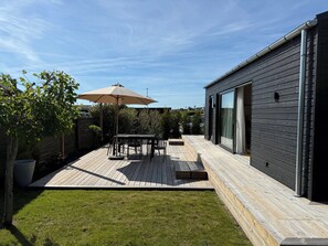 A modern patio with a table and chairs, a large umbrella, and a wooden deck. - Charming newly built cottage 300 m from beach (Karrebæksminde)