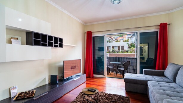 Ein modernes Wohnzimmer mit Flachbildfernseher, einer Couch, einem Couchtisch und einem Balkon mit roten Vorhängen.